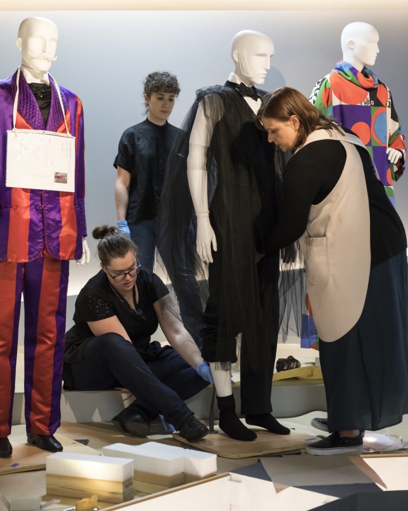 Megan Hall, Conservator (MAAS), Jessica McLean, Registrar (MAAS) and Melinda Kerstein, Costume Installation Specialist (LACMA) securing a half-dressed mannequin to the plinth